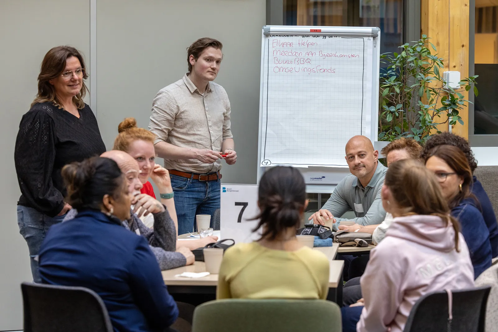 Groep inwoners met elkaar in gesprek. Op het bord staat de tekst: "Elkaar helpen, Meedoen aan bijeenkomsten, BuurtBBQ en Omgevingsfonds"
